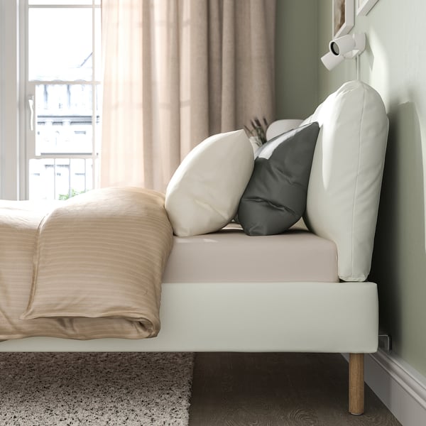 Modern bed frame with sloped headboard adorned with two pillows, positioned against a window with beige curtains. The frame is white with wooden legs.