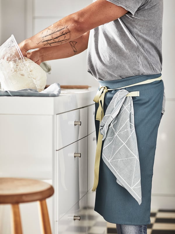 A person in a grey shirt bakes bread wearing a dark blue apron with a white geometric pocket and yellow ties, set in a white kitchen.