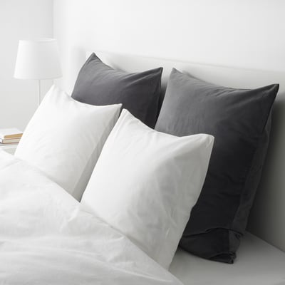 Four square pillows: two white, two grey. One leans against headboard with zip for easy removal. Neatly made bed with white sheets.