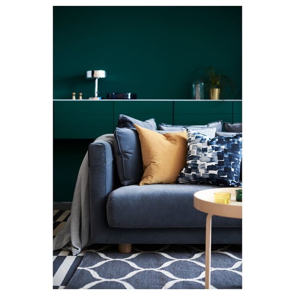 A stylish living room featuring a grey SANELA velvet cushion on a matching sofa. The room has teal walls, modern decor, and patterned rug.