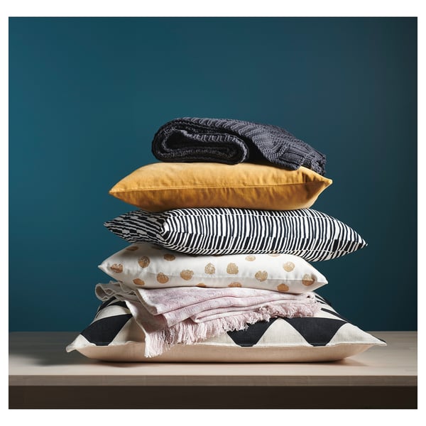 Stacked colourful pillows, mustard, striped, dotted, zigzag patterned on wooden shelf, black wall.