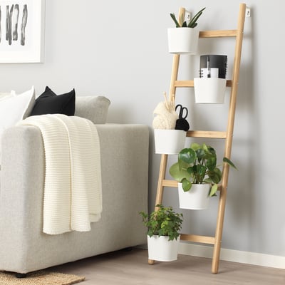 A bamboo ladder plant stand with white pots, beside a grey couch and white blanket.