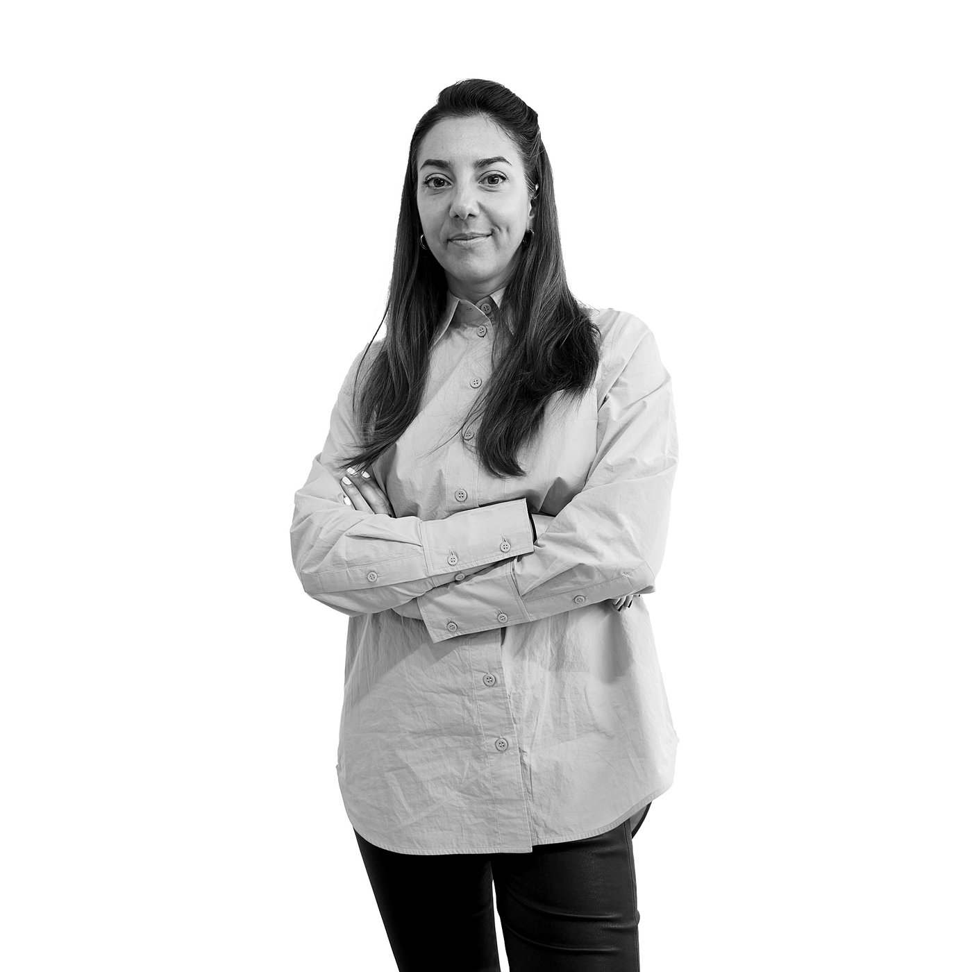 Black and white portrait of the smiling designer with long hair, wearing a button-up shirt and dark trousers, posing with arms crossed.