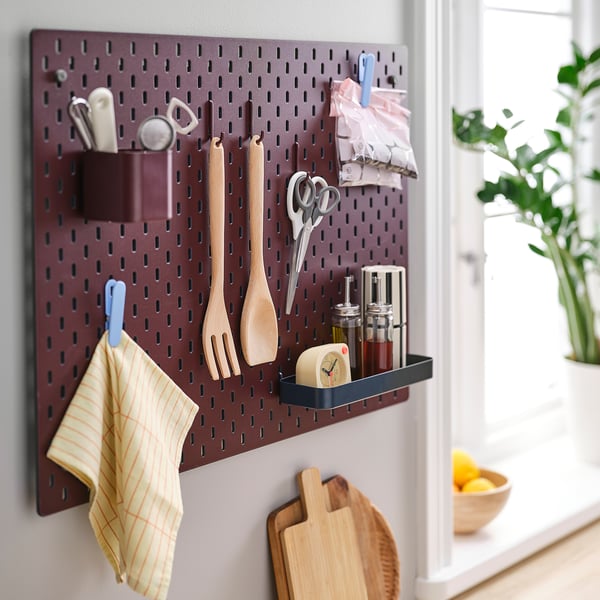 Burgundy SKÅDIS pegboard with hooks, shelf, scissors, wooden utensils, cloth, and cutting board. Organises kitchen tools neatly.