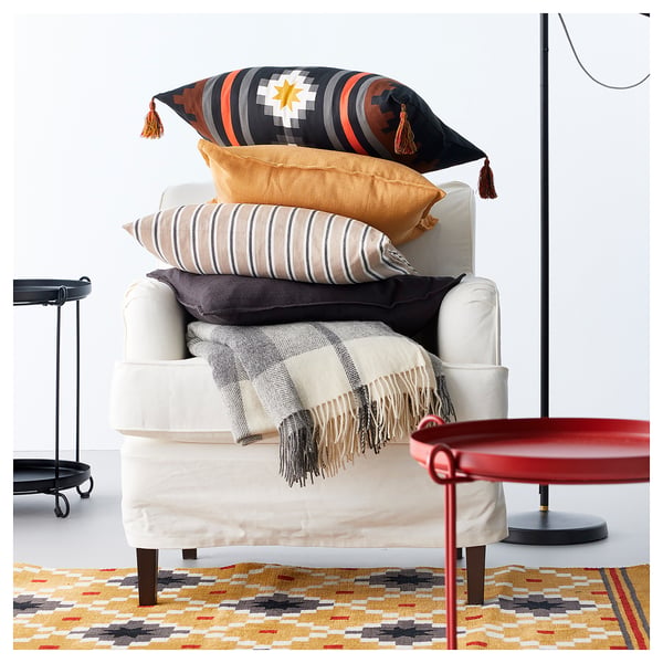 Bright living room scene with off-white armchair piled with colourful pillows and blanket, patterned rug, and modern side tables.