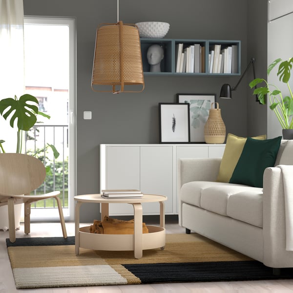 Modern living room with SKRIFTSPRÅK rug in earthy tones, white coffee table, grey armchair with colourful pillows, light wood chair, and hanging lamp.
