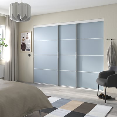 Modern bedroom with grey SKYTTA wardrobe. It has sliding doors, a rug, and a bed with a beige blanket. A black chair sits by the window.