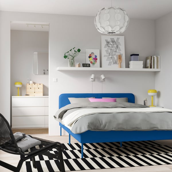 Modern bedroom with blue bed, yellow lamps, black&white rug, shelves, pendant light, dresser, and chair with blanket.