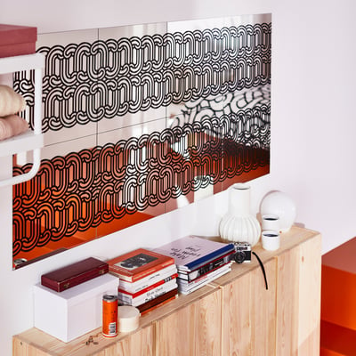 Black & white SÖTRÖNN mirror blakes gallery wall, reflecting items, beside light wood cabinet with books, vases, camera.
