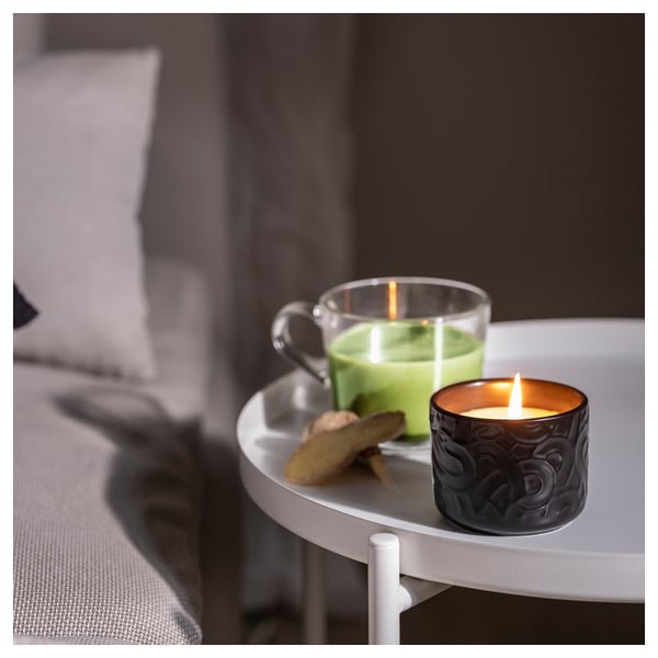 Candle with green tea scent on small table, beside glass mug and biscuits.
