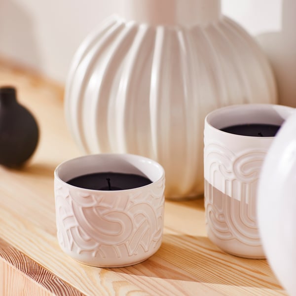 Three white SÖTRÖNN candles. Two small, textured ceramic cups, one large ribbed vase. Small candles fit in hand, vase larger on table.