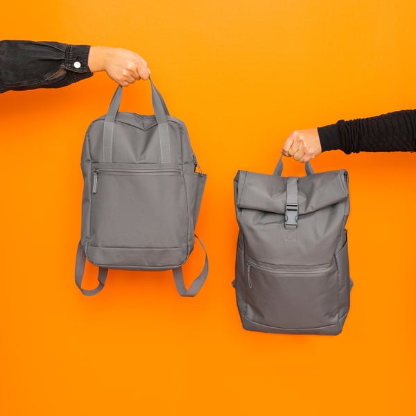 Two hands hold grey STARTTID backpacks against orange background, showcasing water-resistant fabric, outer compartments, and padded shoulder straps.