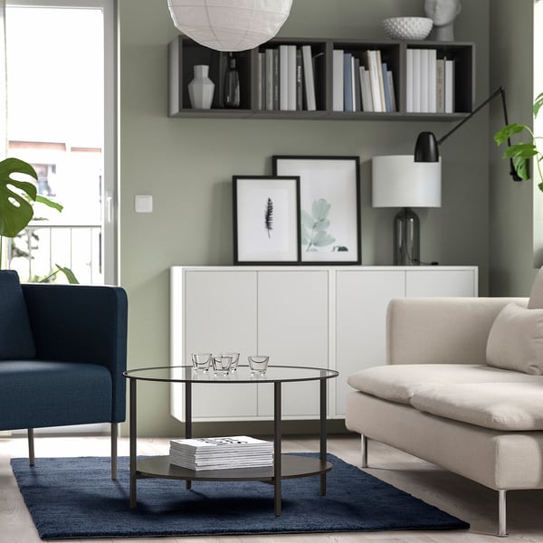 Modern living room with green walls, white sofa, blue chair, dark rug, glass table, art, lamp, books, plants.