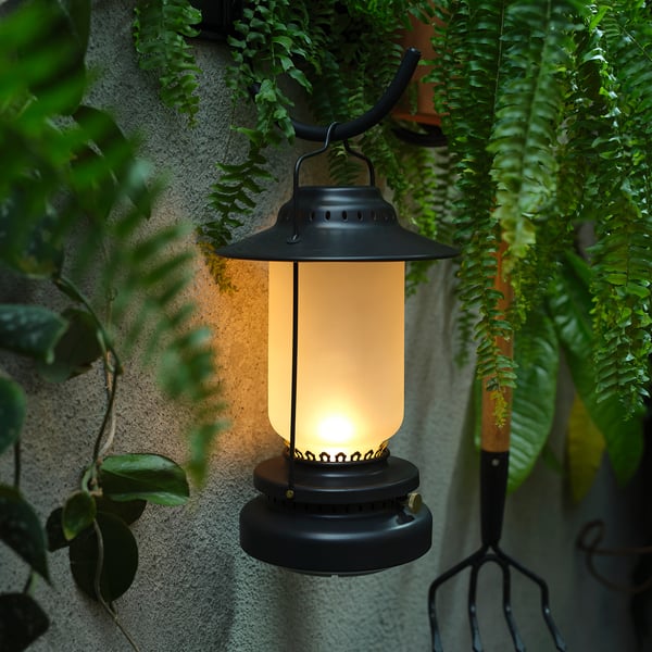 Black STORHAGA lantern hangs outdoors, emitting warm light, surrounded by green plants.