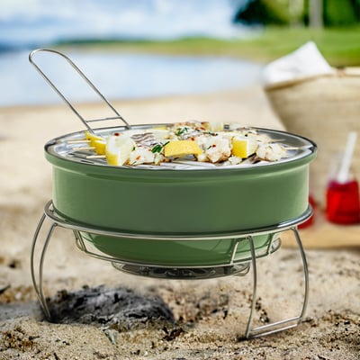 The STRANDÖN grill in green, small and portable, sits on sand with food. Its metal handles and stand elevate it, making it ready for outdoor use.