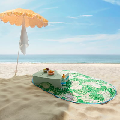 Green picnic blanket with leaf pattern on beach, orange parasol, cooler.