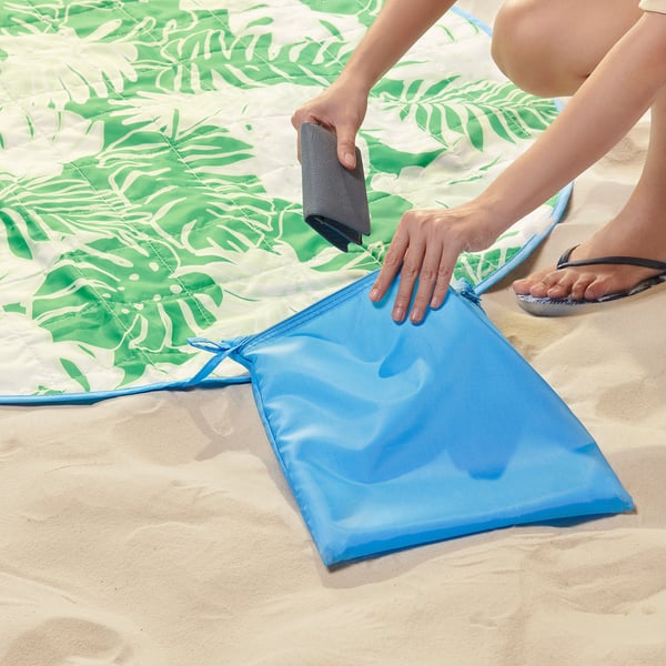 Person placing a phone in a blue pouch of a tropical-patterned STRANDÖN picnic blanket on sand.