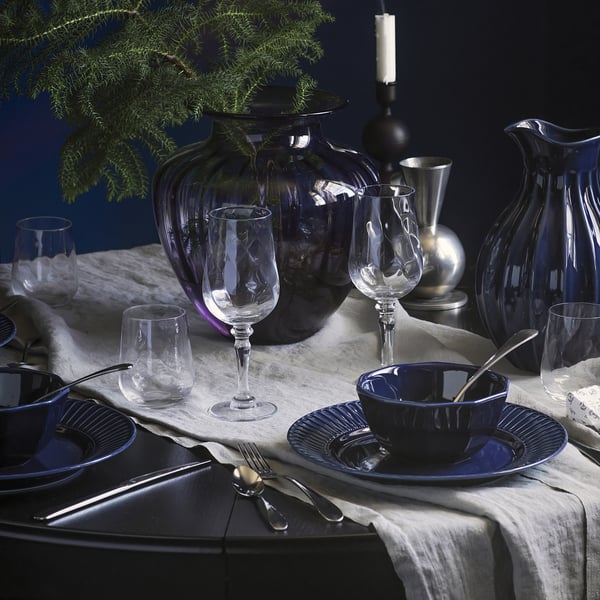 Elegant dinner setup with blue and clear glassware, vases, and dining ware arranged on a white tablecloth. Candles and greenery decorate the table.