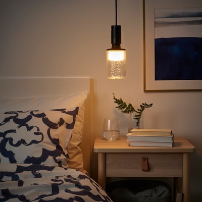 Modern bedroom with SUNNEBY pendant lamp over wooden bedside table, soft glow, patterned sheets, books, and greenery.