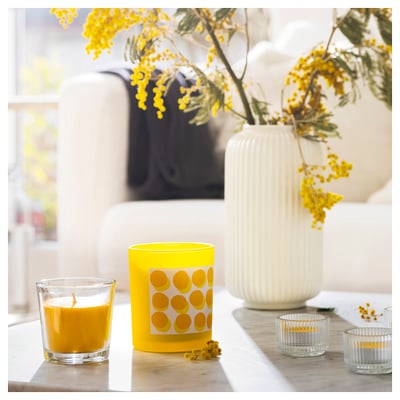 Bright yellow candle in glass holder with polka dots and two clear glass tealights, flowers in white vase on a light wooden table.
