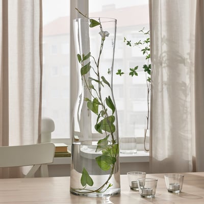Clear glass vase on table near window, holding long stemmed flowers, with smaller glasses beside.