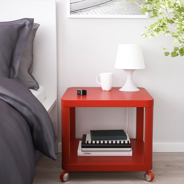 Red TINGBY bedside table with wheels, next to bed. Rectangular with shelf, lamp, mug, and clock.