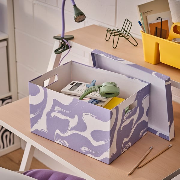 Office desk with purple TJENA storage boxes, containing stationery. Bright yellow container, lamp, and notes. White brick wall background.