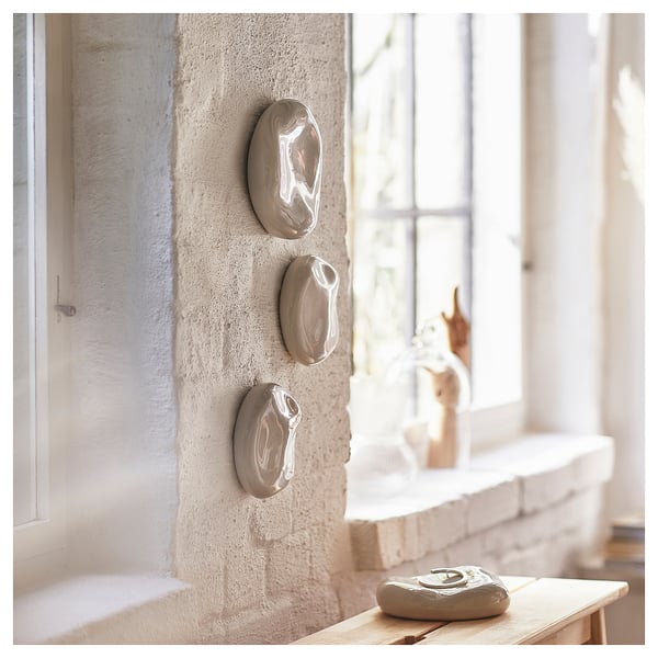Three beige ceramic wall hangings, organic shape, glossy, on textured wall by window with sculpture.