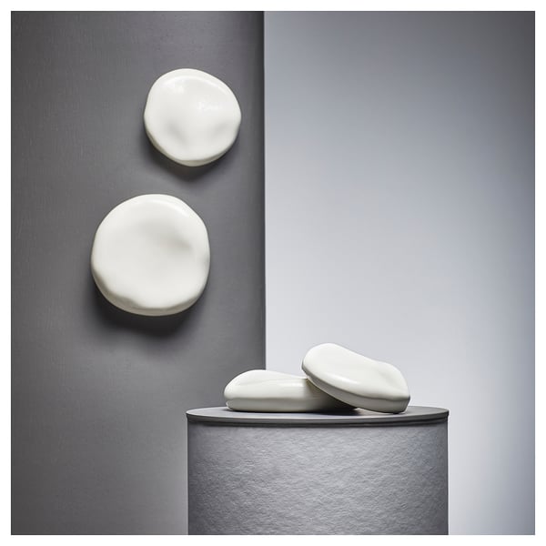 Three white, round, ceramic wall decorations arranged in a minimalist manner, two mounted on a grey wall, one resting on a cylindrical table.
