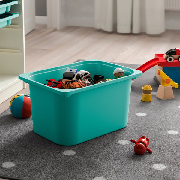Turquoise TROFAST storage bin filled with toys on a grey carpet.