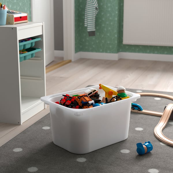 A white plastic storage bin, filled with colourful kids toys, sits on a grey carpet near a toy train track.