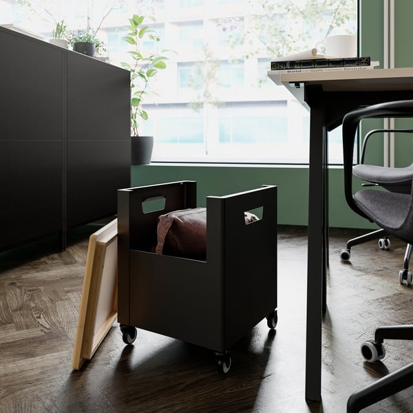 Modern office space black TROTTEN storage on wheels near a window. Contains a bag, with a natural bamboo lid, offering practical storage.