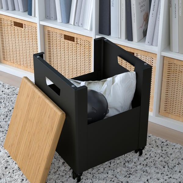 Black storage bin with bamboo lid and wheels on a rug, next to a shelf with books and baskets, holding a white bag.