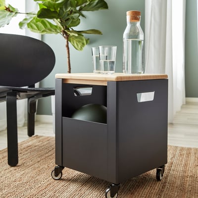 Small black storage box on wheels with bamboo lid. Holds items like bags or binders. Glasses and bottle on top.