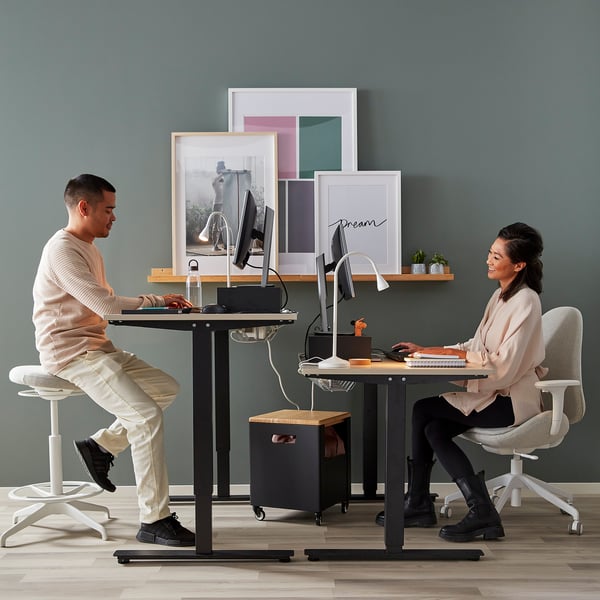 Two people working at standing desks with a storage unit between them. One with a laptop, the other on a computer. Art and decor on the desk and wall.