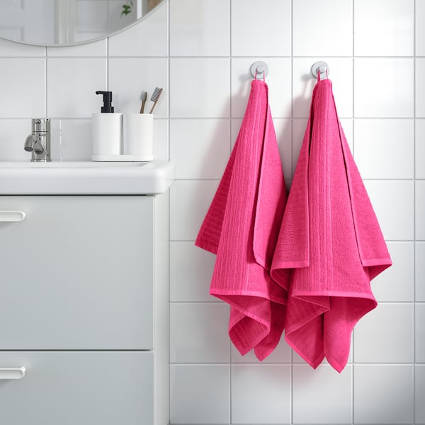 Bathroom with bright pink VÅGSJÖN towels hanging, white wall tiles, sink with soap dispenser and toothbrushes, cabinet below.