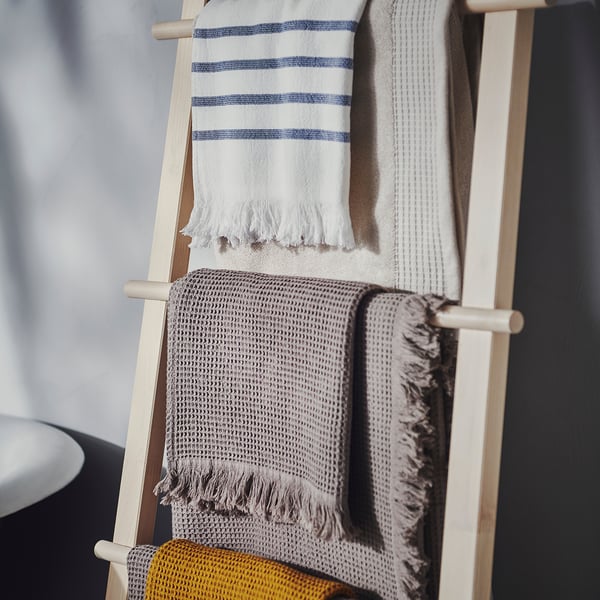 Wooden towel rack with three towels. Top towel: light grey with blue stripes. Middle towel: grey with fringe. Bottom towel: yellow and grey weaved.