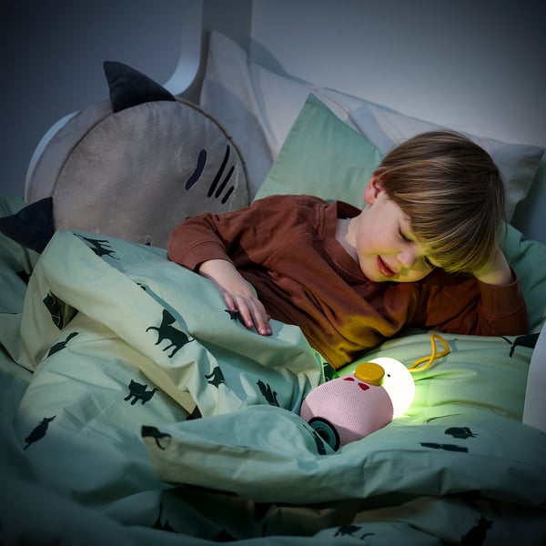 Child lays in bed, cuddling a toy, beside a glowing speaker lamp, with a plush shark and cat-patterned blanket.