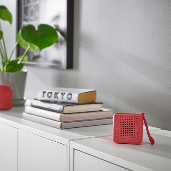 Small red VAPPEBY speaker on shelf, near books, plant, and art. Compact, perforated design.