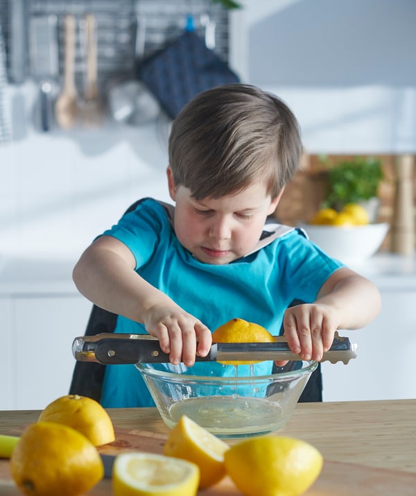 A young boy in a blue shirt squeezes lemon onto a juicer in a kitchen. A glass bowl catches the juice on a wooden countertop.