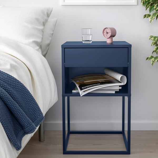 Blue bedside table with drawer and shelf holding glass, clock, and magazines next to bed.