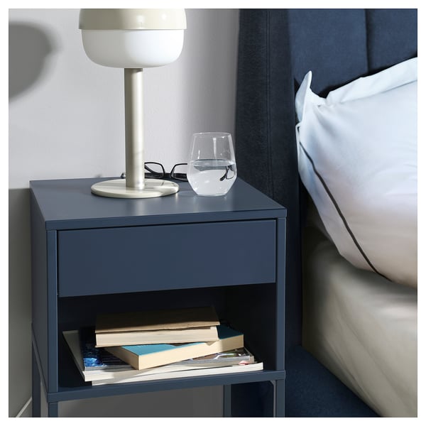 Blue bedside table with white lamp, glass of water, and books next to a bed.