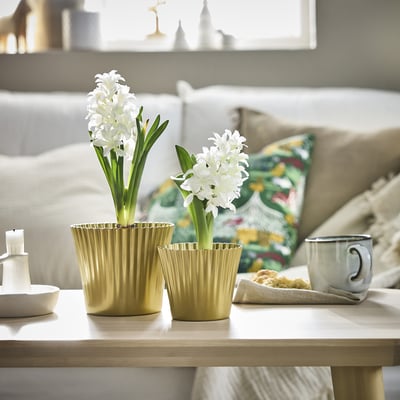 Two VINTERFINT gold plant pots hold white flowers, larger than a mug and a candle, on a wooden table near a beige couch with colourful pillows.