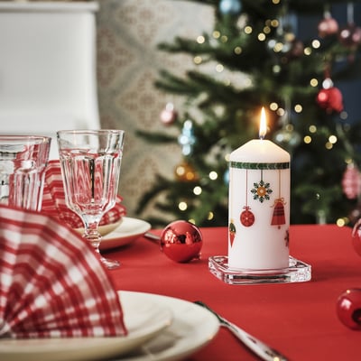 Holiday table setup with VINTERFINT white pillar candle, featuring red decorations. Larger than nearby wine glasses, on red tablecloth.