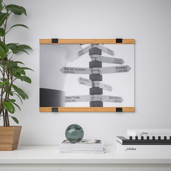 Wall-mounted poster hanger with dual parts holding a directional signpost poster, supported by a wooden shelf with a plant and decorative items.