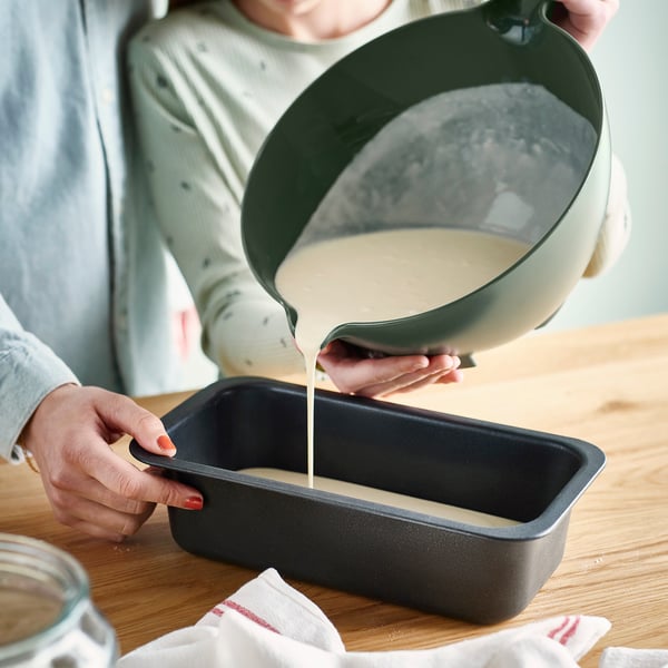 A person pours batter from a VISPNING mixing bowl into a black loaf pan. The bowls handle aids in precise pouring.