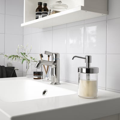 A sleek VOXNAN soap dispenser with a transparent glass mug and chrome details sits on a white countertop in a stylish bathroom.