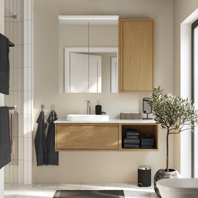 Modern bathroom with ÄNGSJÖN oak vanity, white sink, mirror, dark towels, olive plant. Minimalist decor.