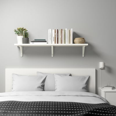 Minimalist bedroom with white BERGSHULT wall shelf holding books & plants above a bed with grey pillows and black patterned blanket.