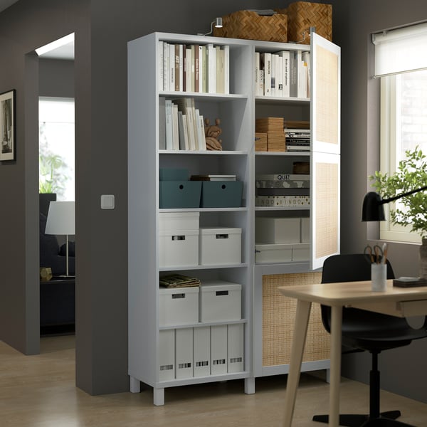 A modern storage unit with open and closed sections, holding books, boxes, and decor, situated beside a work desk with a chair.
